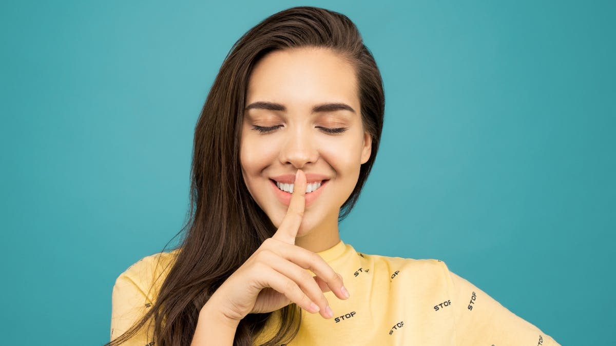 Charming woman with finger on lips, signaling silence against a teal background.